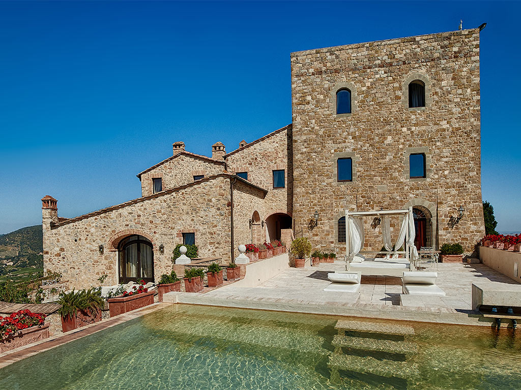 Castello di Velona, em Montalcino, na Itália. É possível ver a piscina, um deck com sofás brancos e o castelo no fundo. 