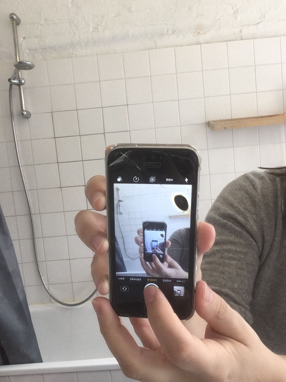 An image of an iPhone taking an photo of itself in a mirror.  White hands hold it up in a bathroom with white tile.