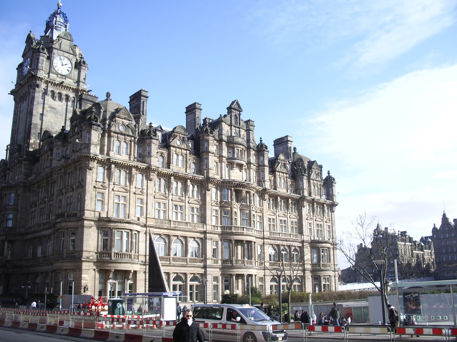 the clock tower of edinburgh. traditional architecture illuminated by the sun on a cold sunny day. car, construction barriers and people at bus stop in front of the clock tower.