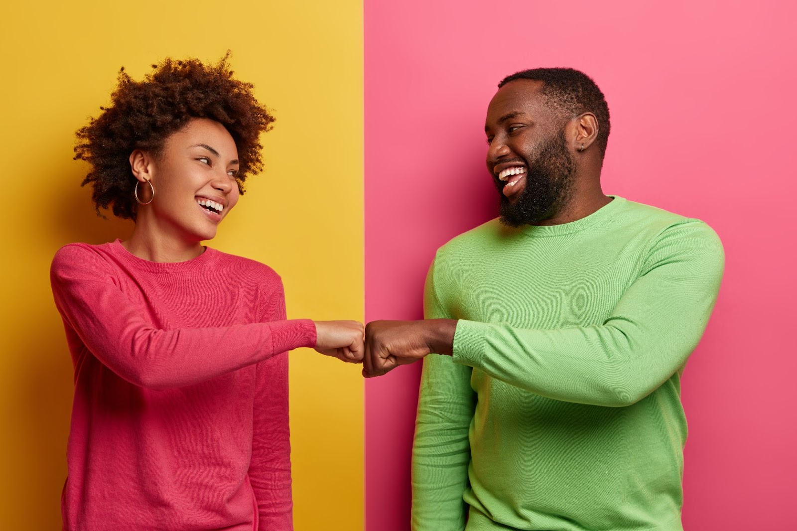 Duas pessoas negras, uma mulher de malha rosa e um homem de malha verde, olham felizes um para o outro e batem os punhos