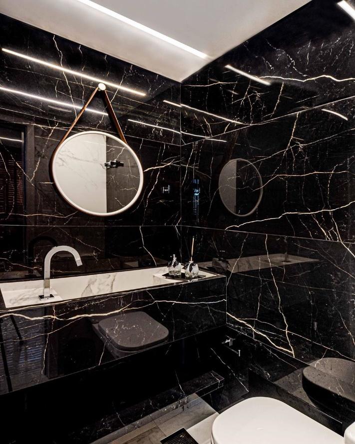 Banheiro todo revestido com porcelanato marmorizado preto, espelho redondo com moldura branca e louças brancas.