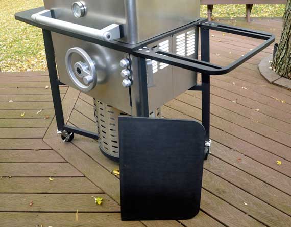 A shiny rectangular gas grill with a removable black side shelf on the ground