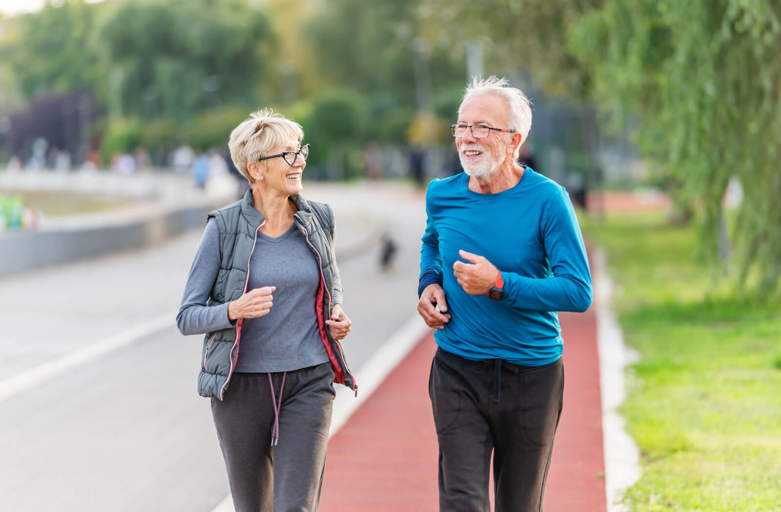 A senior woman and a senior man smiling while jogging in a park.