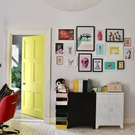 Quarto com porta amarela, piso de madeira branca, parede com muitos quadros coloridos e armários pequeno preto e branco.