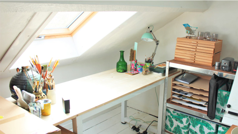 A small attic that’s been converted into a cozy crafting corner. Natural light is pouring in from the skylight window onto the crafting table.