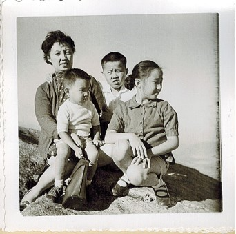 Woman seated at left holds a small child on her lap. Boy in center looking at camera is behind a girl looking to the right.