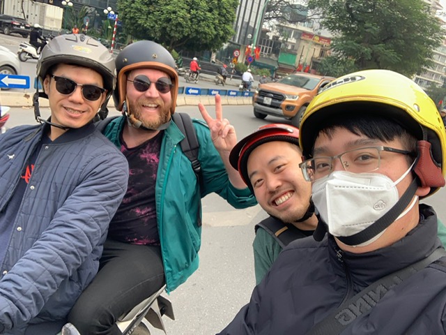 Four guys wearing helmets and sunglasses pose while riding on scooters in the streets of Hanoi Vietnam.
