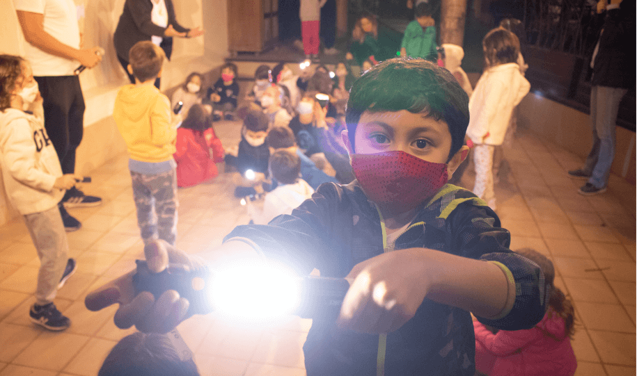 A imagem mostra, em primeiro plano, um menino com uma lanterna; atrás, várias crianças.