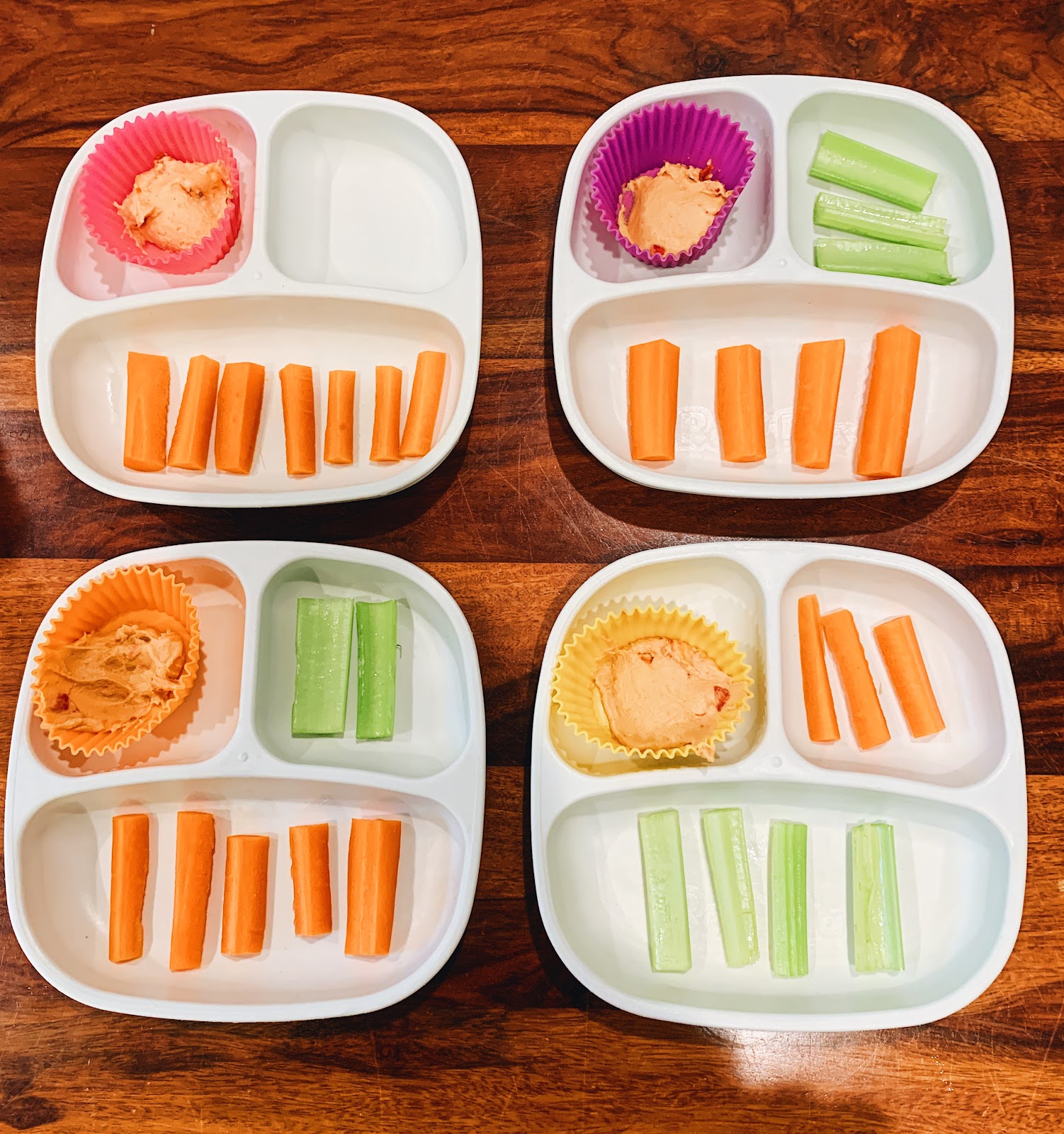 One snack tray has hummus and 7 carrots. No celery. Another tray has hummus, 4 carrots, and 3 celery sticks. A third snack tray has hummus, 5 carrots, and 2 celery sticks. The last tray has hummus, 3 carrots and 4 celery sticks.