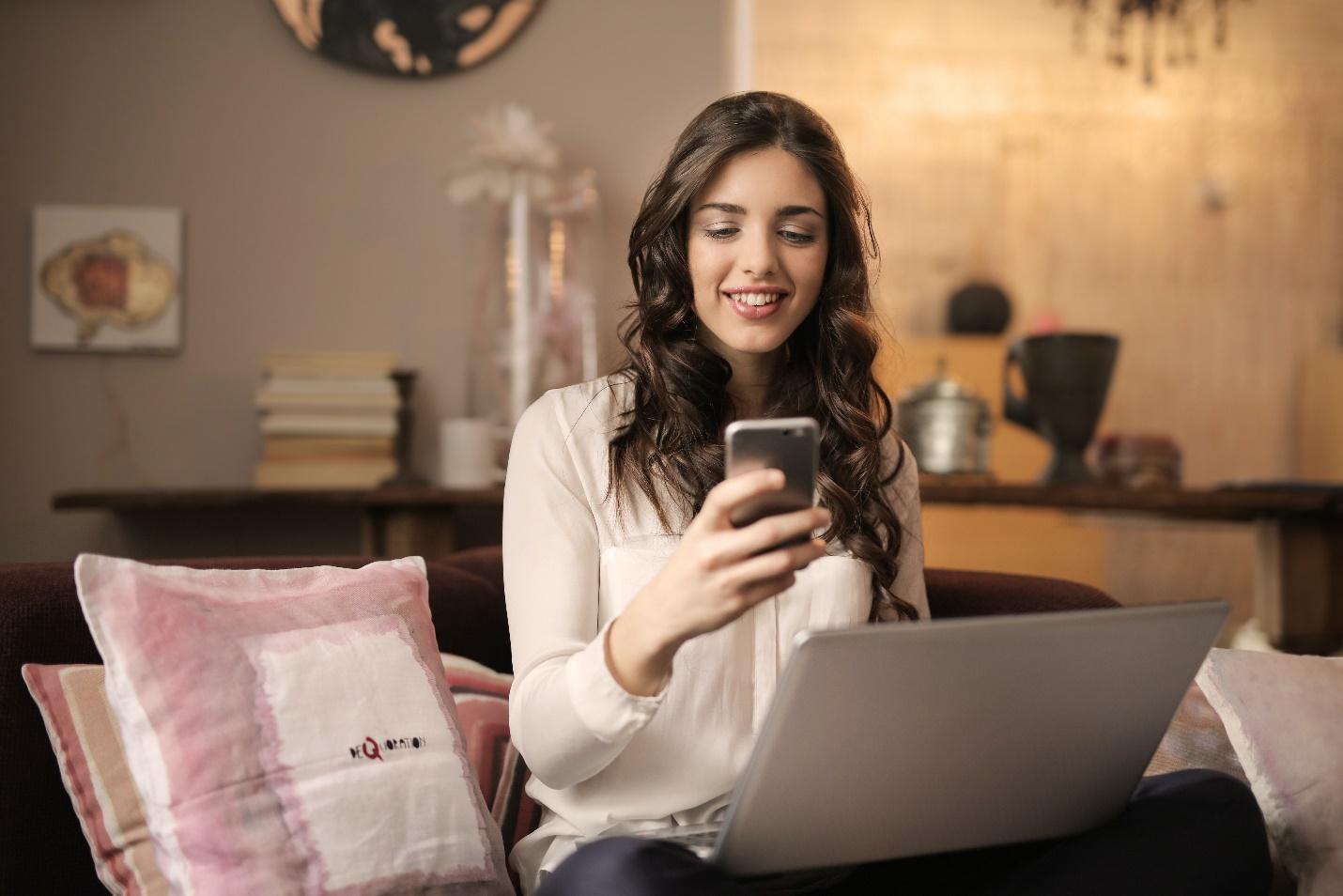 cum sa faci bani online o femeie tanara navigheaza online pe telefonul si si pe laptop stand pe canapea