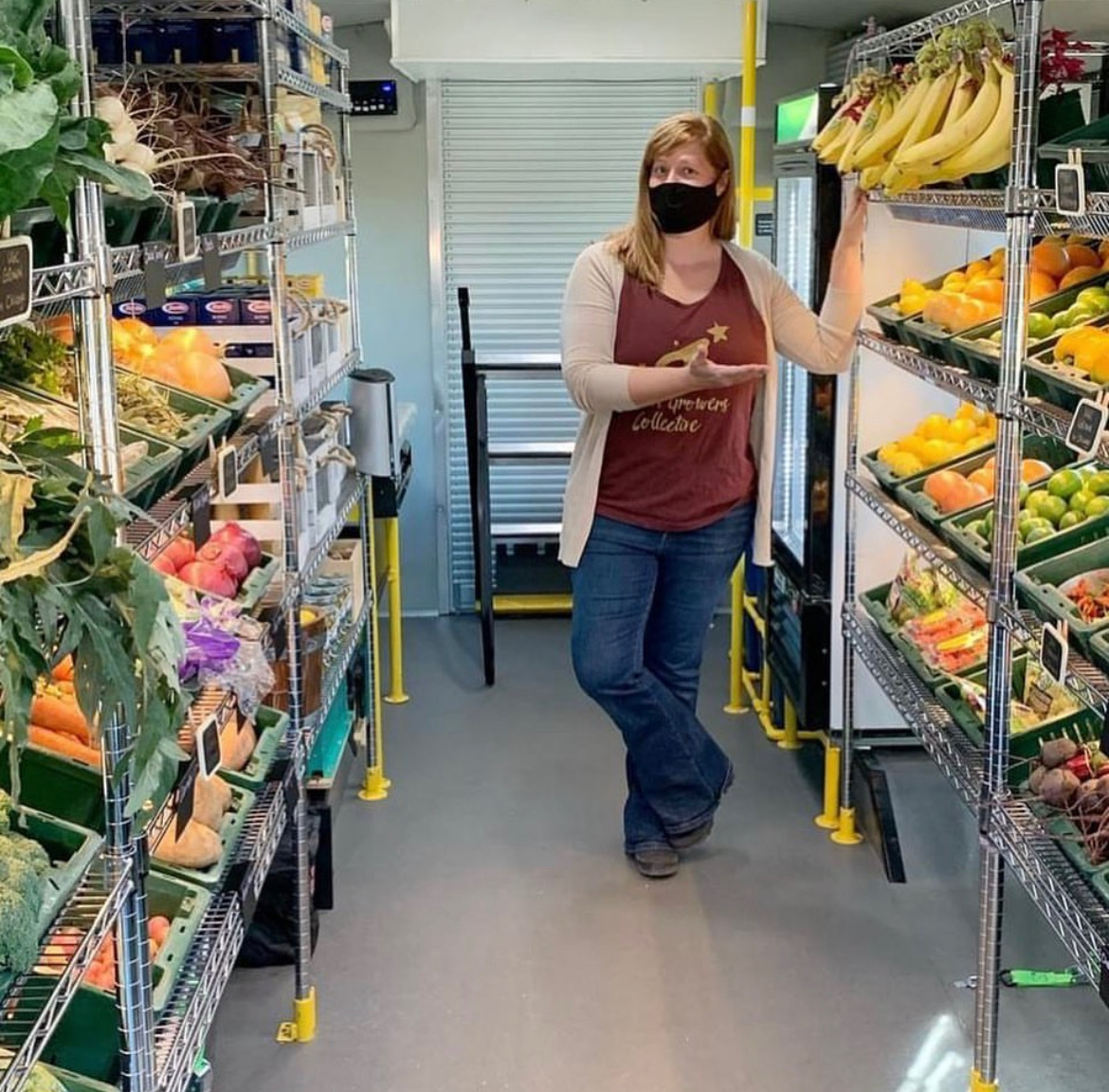 A woman stands between two rows of stocked fruits and vegetables and gestures toward some bananas.