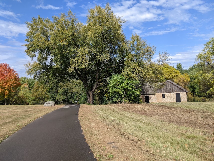 An asphalt trail lined with grass has an orange stone and wooden building with a large green deciduous tree surrounded by red, green, and yellow trees in the background. 