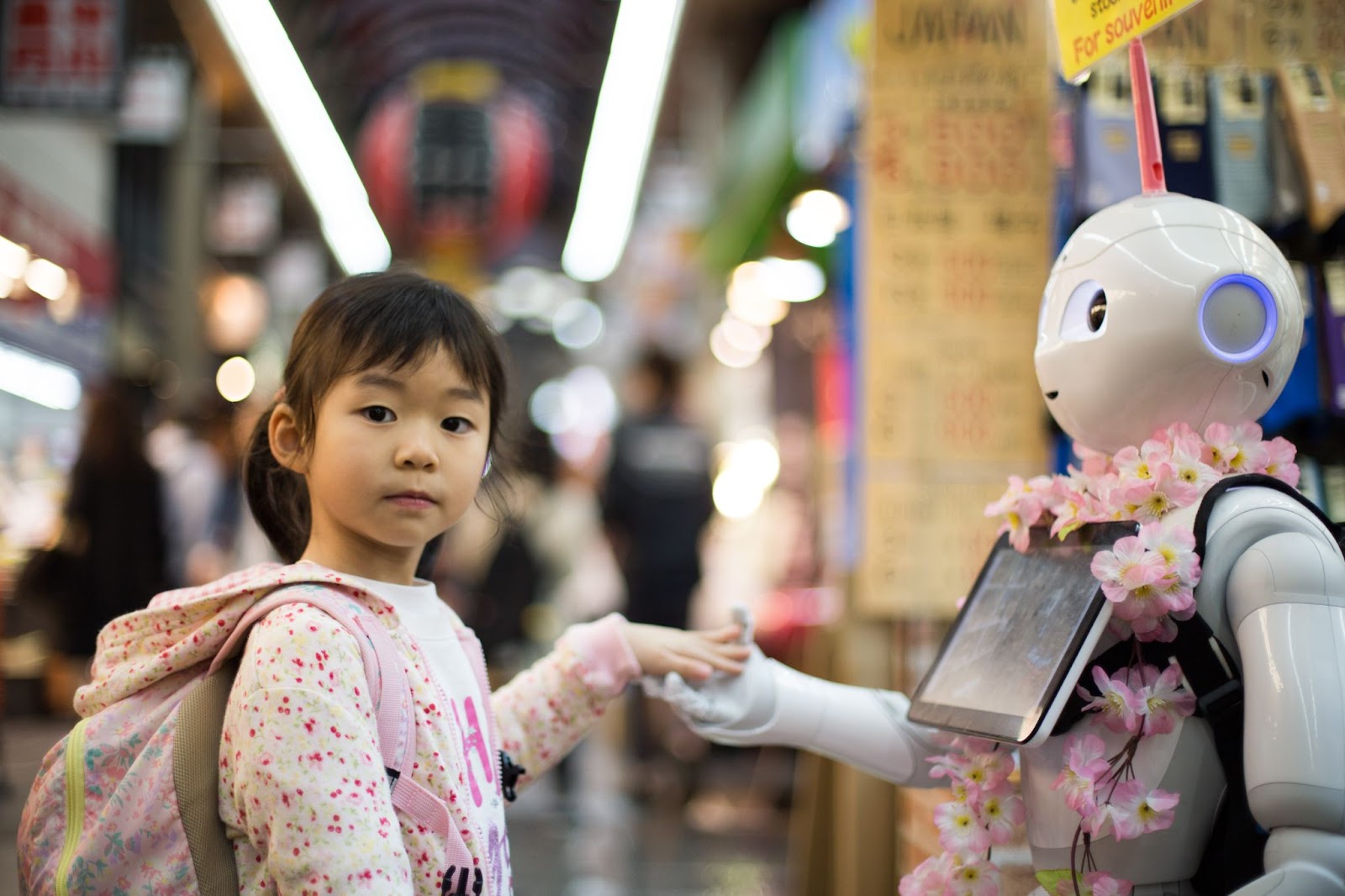 Menina japonesa está em uma feira, segurando a mão de um robô do seu tamanho, para ilustrar uma inteligência artificial. 