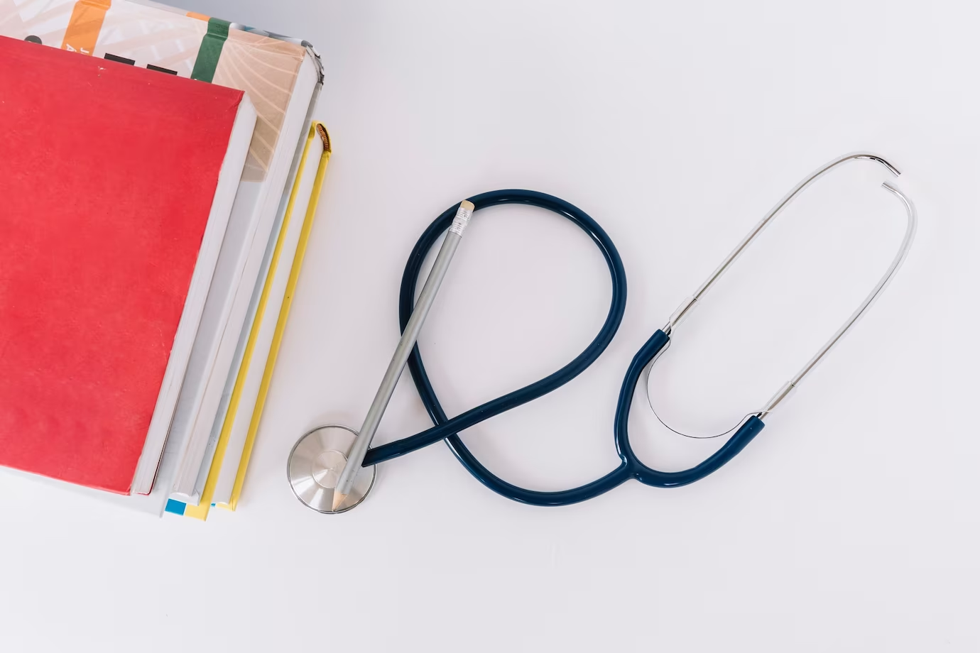 High-angle view of stacked books and a stethoscope on a white surface, representing essential subjects for aspiring doctors in 'Your Roadmap to Medicine.'