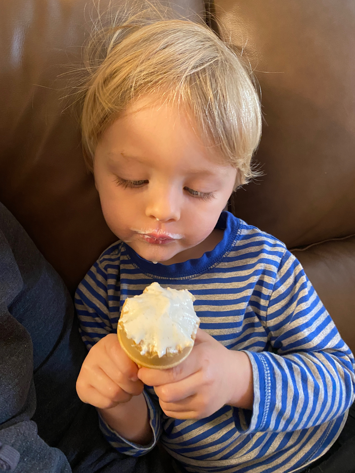 Toddler on the sofa wearing a striped shirt with an ice cream cone in his hands and ice cream on his face