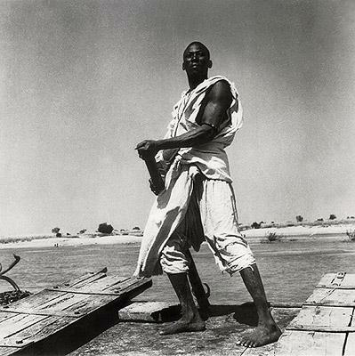 Registro Fotográfico do artista. Um homem negro com vestes brancas, segurando um pedaço de pau. Ele está centralizado um pouco para direita, ao chão de terra algumas madeiras. Ele está com a feição séria e dirige o olhar para frente.