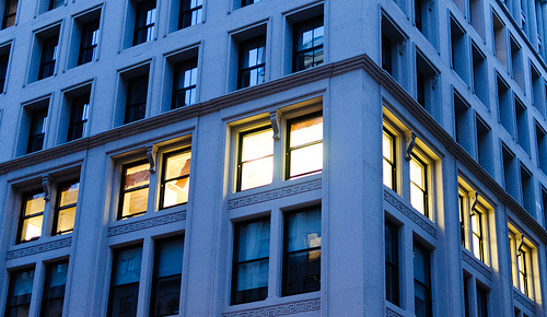 Tall building with lots of windows on each story. The camera angle is a the corner of the building looking upwards slightly. The lights on the second floor are all on while the rest of the stories have their lights off.