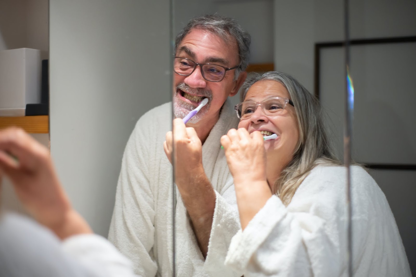 A imagem mostra um casal de idosos escovando os dentes juntos em frente a um espelho, enquanto vestem roupões de banho. Eles estão dentro de um banheiro.