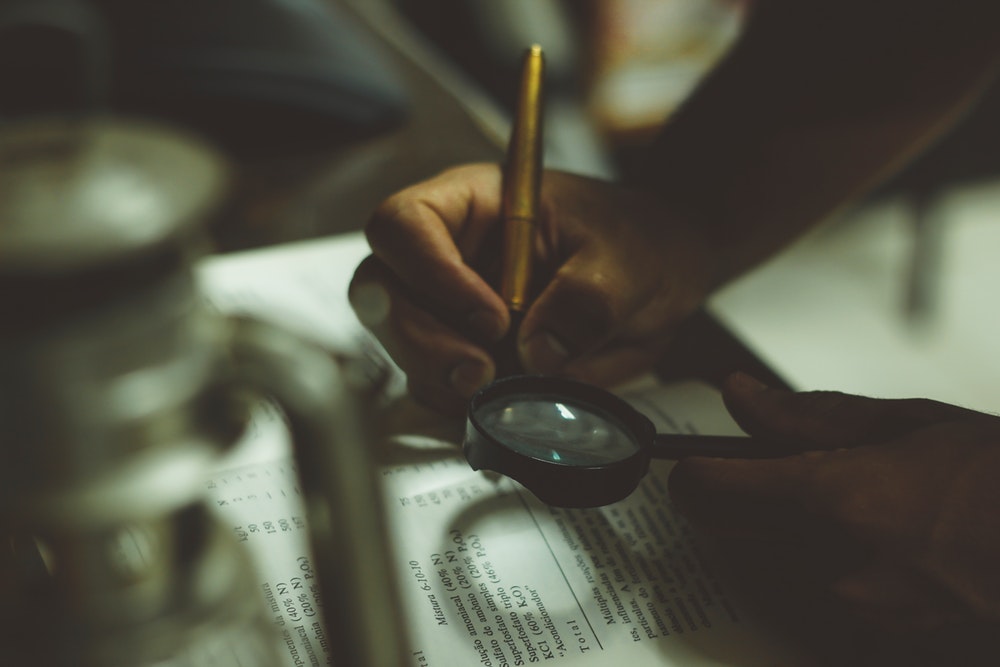 A person reading a book with a magnifying glass and a pen in hand