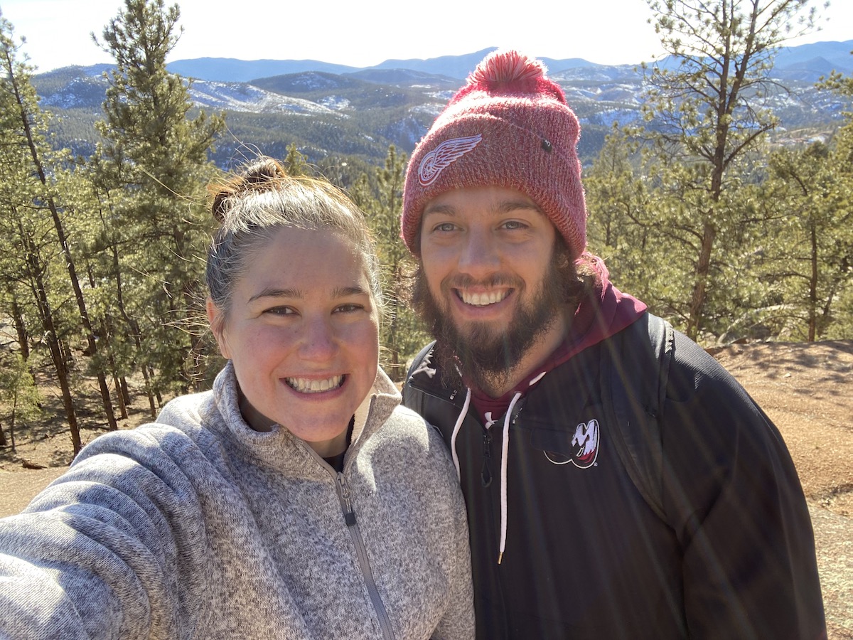 Allie from the Hoppy Passport is standing with her husband  mountainside overlooking the forest and a mountain range. Allie is wearing a white patterned jacket. Her husband is wearing a red beanie/toque and black jacket.