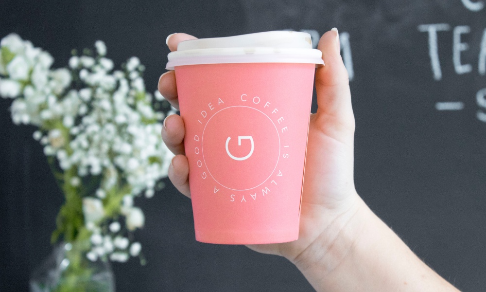 Female Caucasian hand holding double wall paper takeaway coffee cup with pink and white label with white lid.