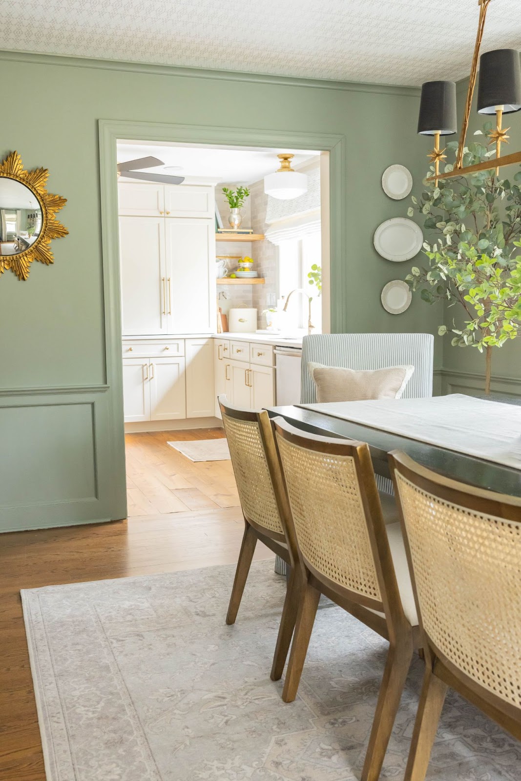 A well-designed dining room features a soft green wall and rattan midcentury dining chairs. The room's elegance is enhanced with a gilded mirror and white porcelain plates displayed as wall decor.