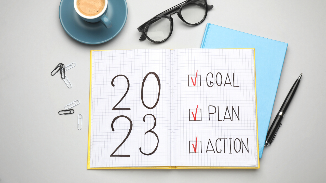 A notebook with 2023 goal plan action written inside it on a desk with glasses, coffee and office supplies