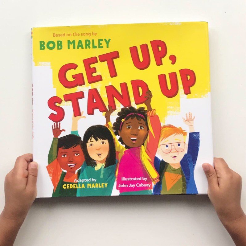 Book cover of Get Up, Stand Up by Bob Marley and Cedella Marley; children's hands holding the book, four cartoon children raising their hands