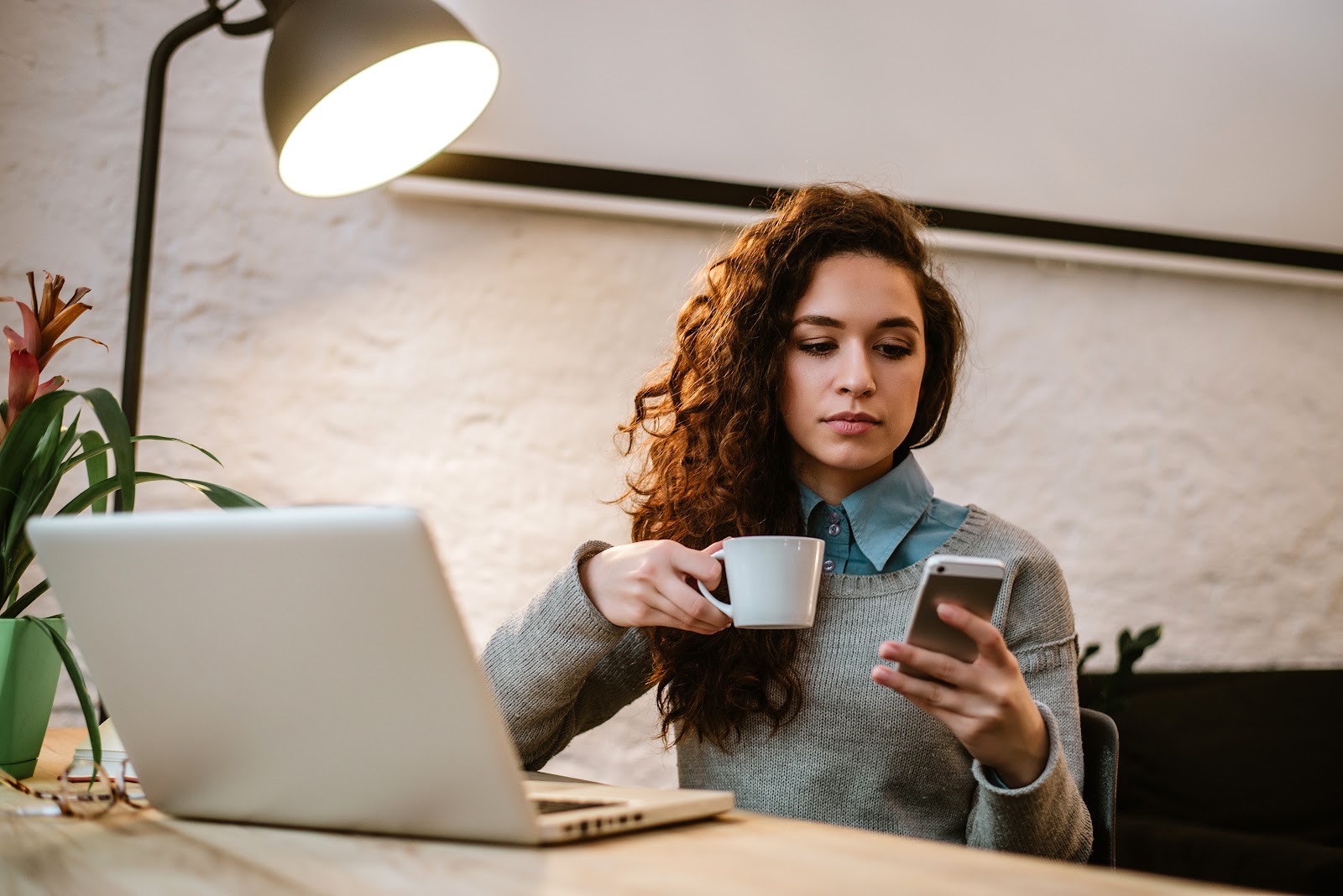 Young woman drinking coffee at her desk while looking at her smartphone