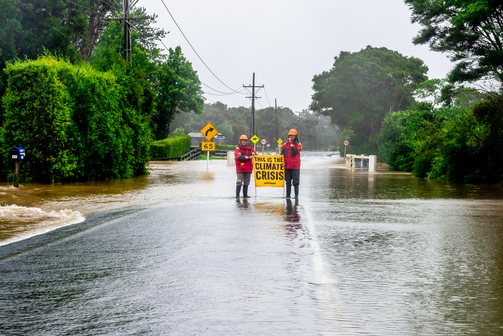 A road underwater, two people in red jackets and hard hats hold a banner that says This is the climate crisis