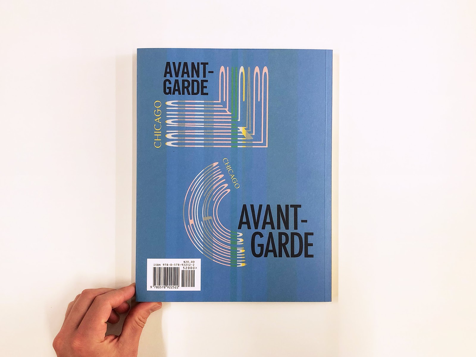 Image: The back cover of the catalog. A matte blue cover, with the bottom lefthand corner held by a hand. On the cover are the words in black "AVANT-GARDE" in the top left corner and the bottom left corner, both framed by distinct words: "CHICAGO," pink and stretched. Photo courtesy of Sonnenzimmer.