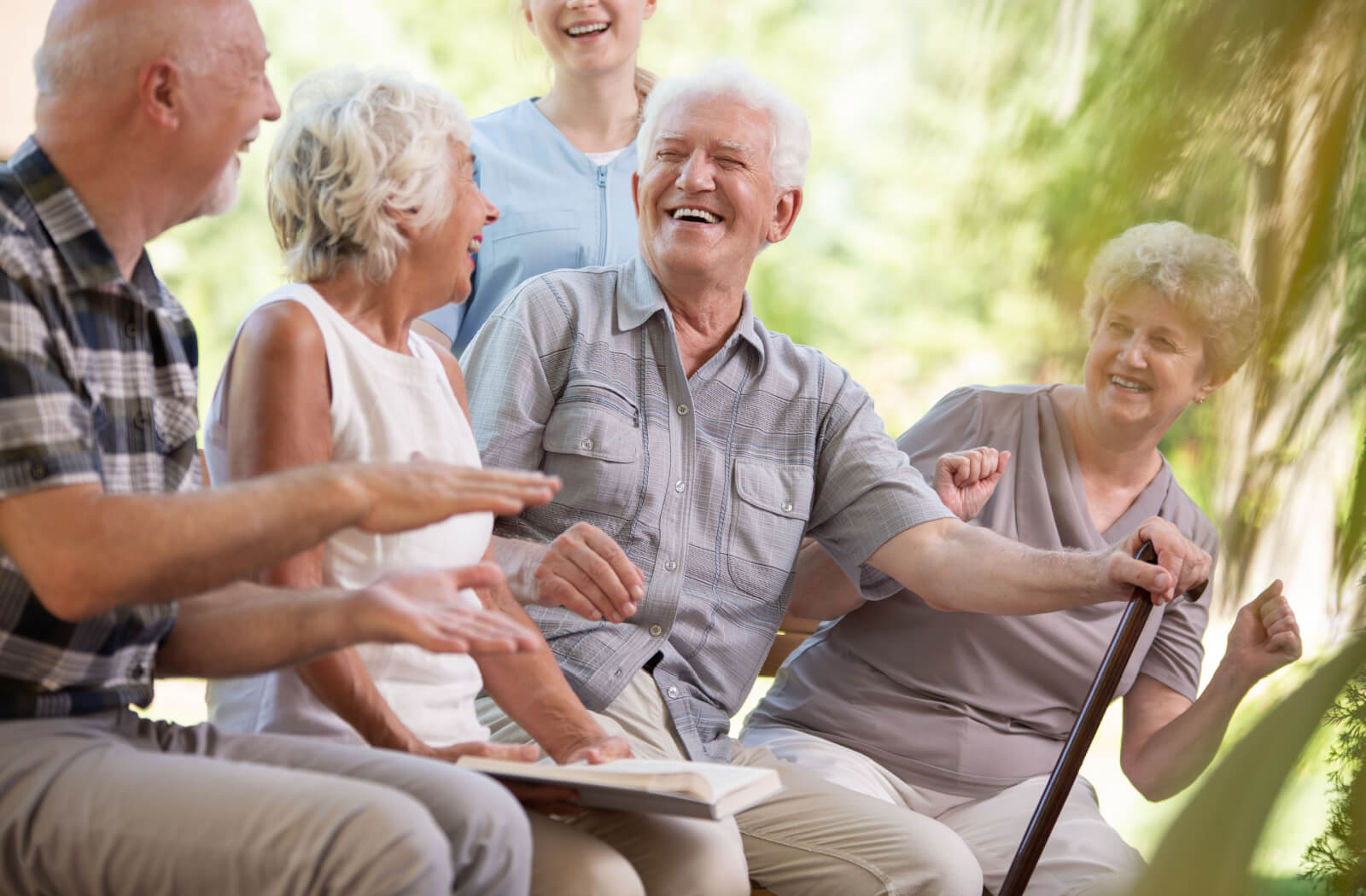 A senior man with a cane smiles and laughs while sitting outside with a nurse and other seniors.