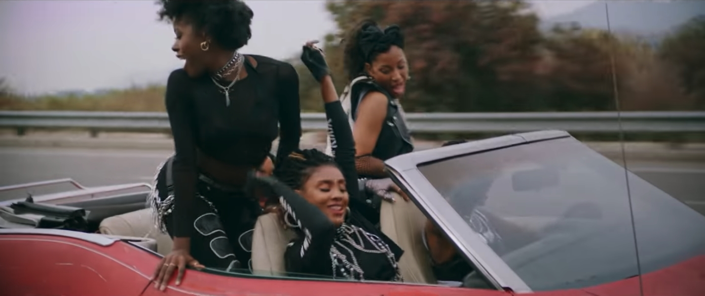 Black women dancing in their seats, standing up in their seats of the red convertible, as it drives. They look happy.