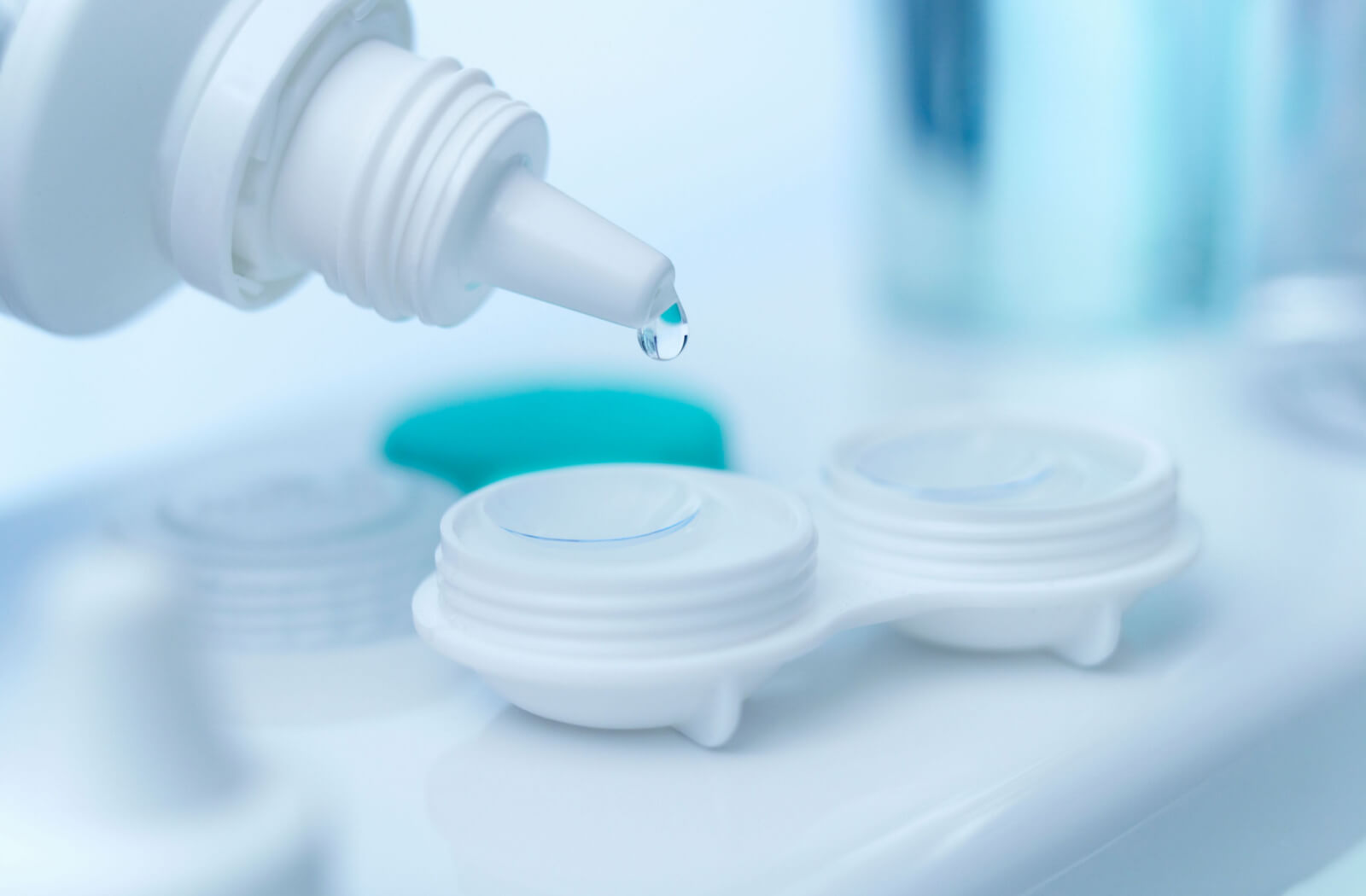 A photo of a white-colored contact lens case with the contact lenses inside and a bottle of solution being poured into the lenses for storage.