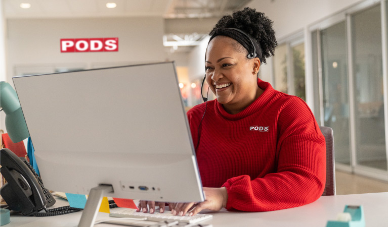 Penny, a PODS customer service rep, smiles at her desktop computer as she helps a PODS customer plan their move over the phone.