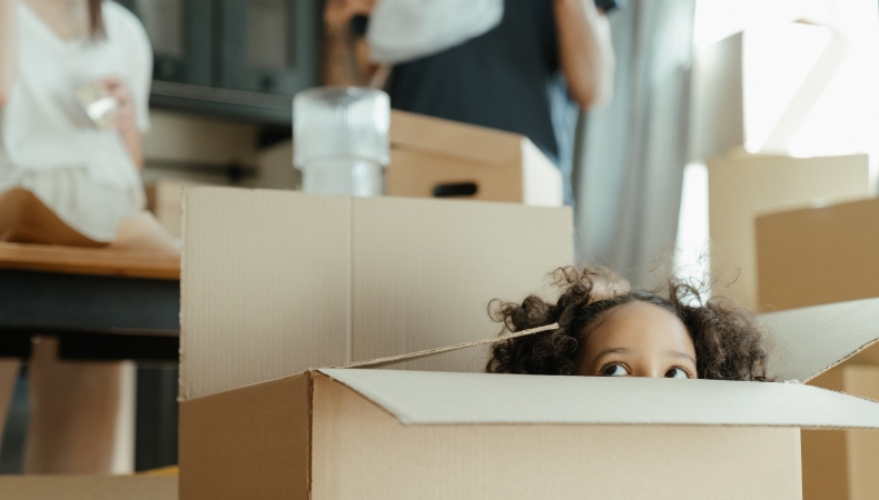 A little girl is peeking out of a moving box as her parents are packing up dishes in the kitchen behind her.