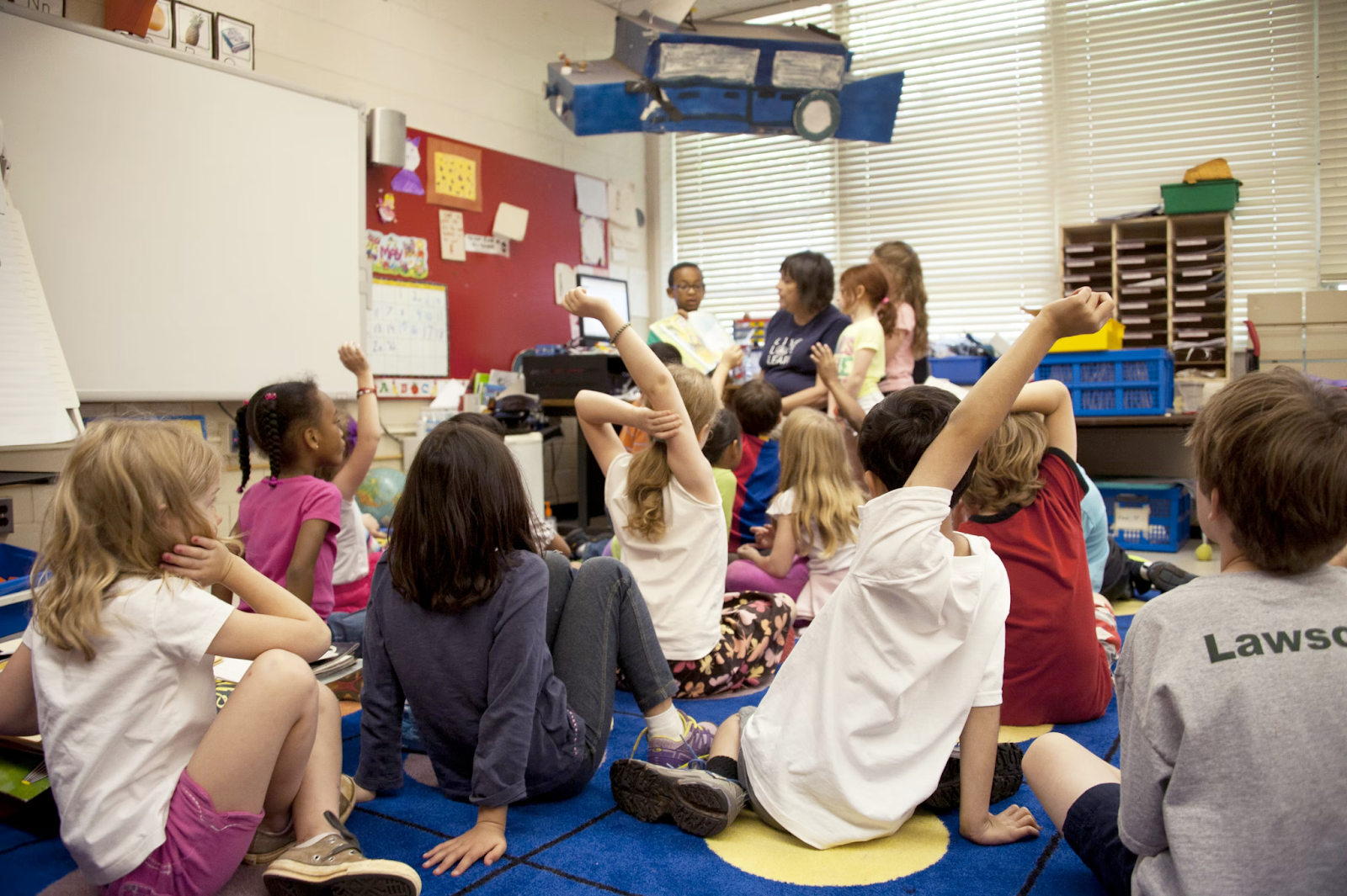 Children sitting together in a classroom. Some children have their hands up to participate.