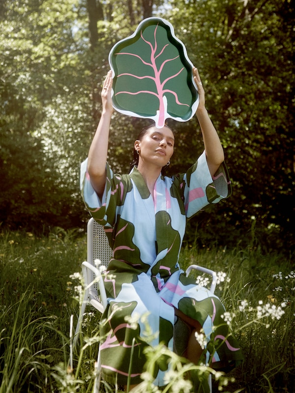 A woman wearing a BASTUA kimono with a leaf pattern sits on a chair in a meadow while holding up a BASTUA leaf-shaped tray.
