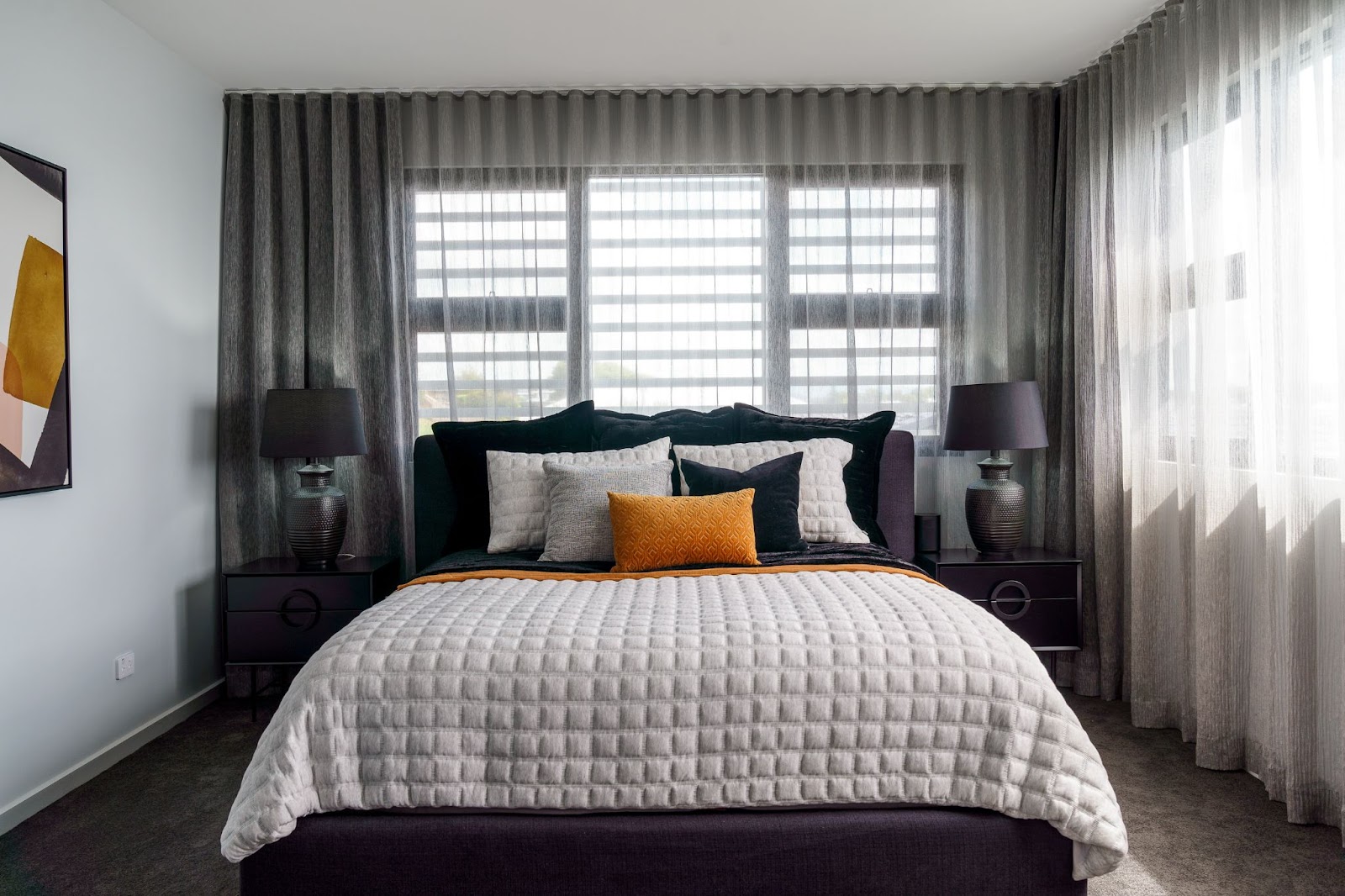 A cozy bedroom with a large bed featuring a tufted white duvet, black and gray pillows, and a contrasting orange accent pillow. The room is softly lit by natural light filtering through sheer curtains, with matching bedside tables and lamps on either side.