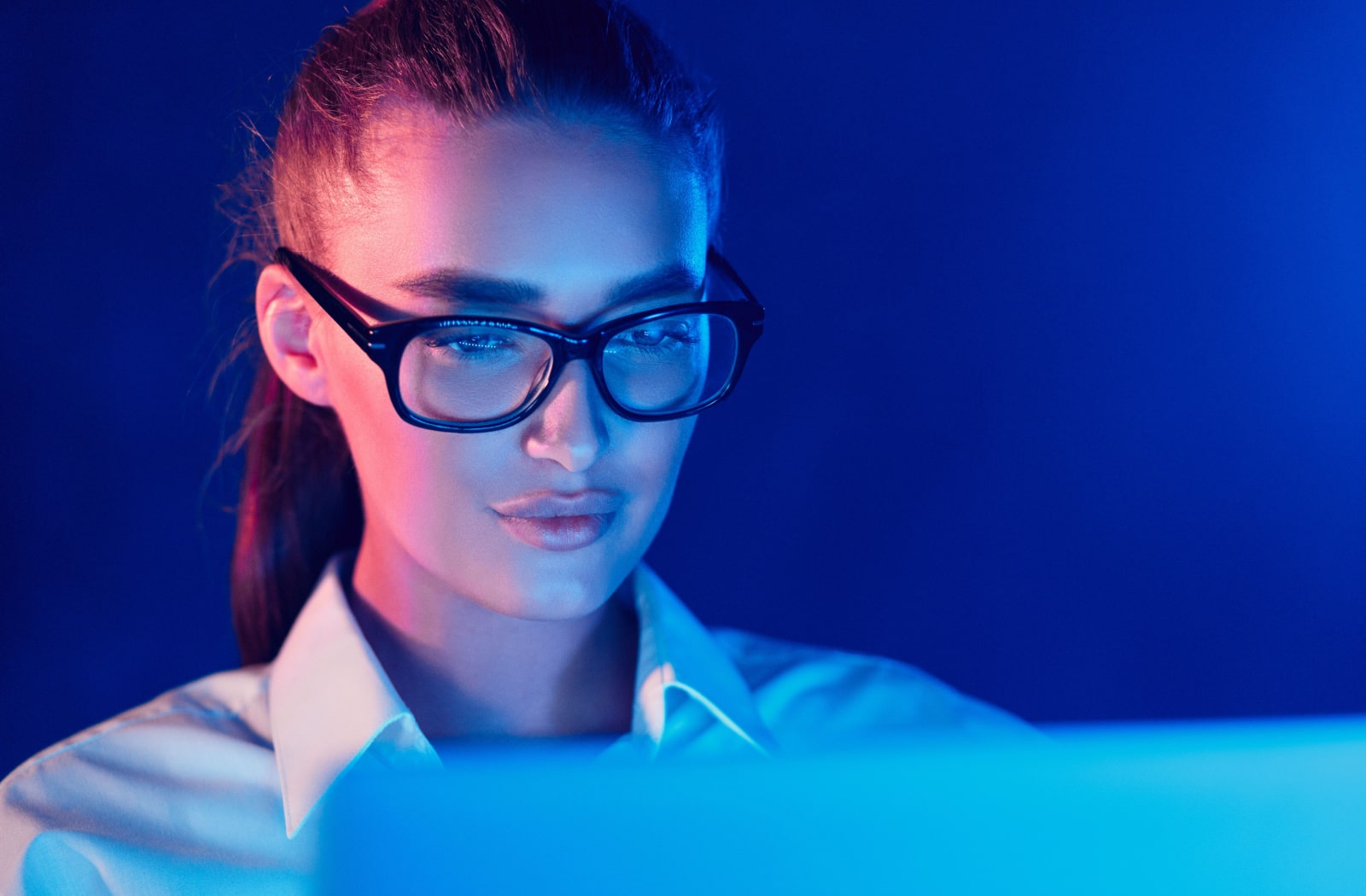 A young woman wearing blue light protection glasses while working on her laptop at night.