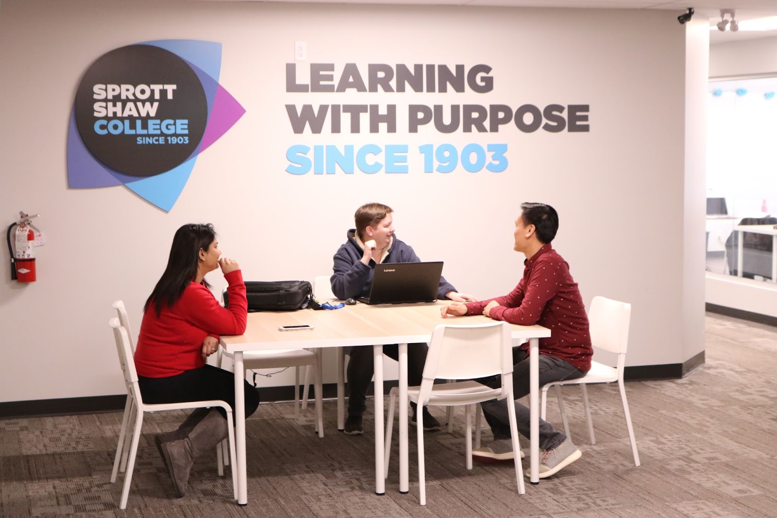 a study area in Kelowna's Sprott Shaw College, with three students sitting at a table working on laptops and a logo sign on the wall