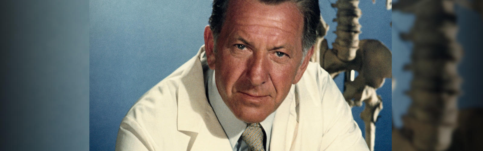 Jack Klugman as Quincy, M.E., looks at the camera with anatomical skeleton in the background
