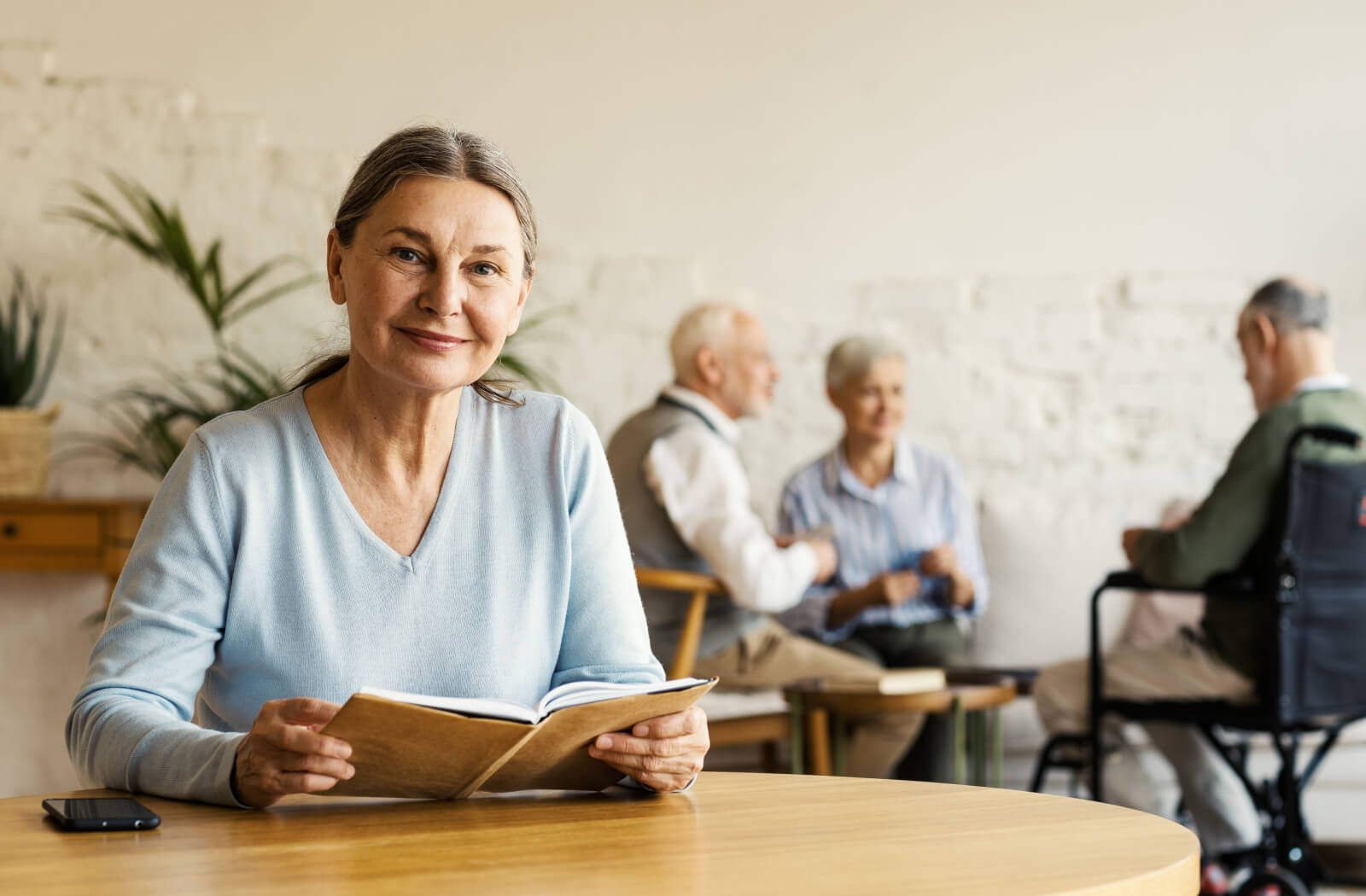 A senior woman smiling while sitting at a table holding a book with other seniors in the background.