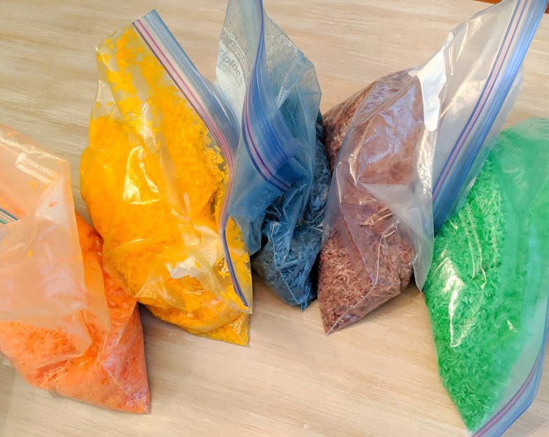 Five different bags of rice dyed with food coloring in gallon Ziploc Bags. The colored are orange, yellow, blue, purple, green. 