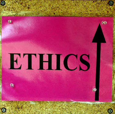 A bright pink sign with Ethics and an upward-facing arrow, tacked on a corkboard.