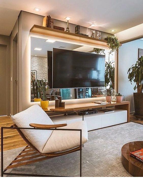 Sala com Painel de TV de espelho, rack de madeira, poltrona, plantas compondo a decoração e piso de madeira com tapete cinza.