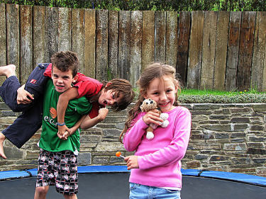 Three children, two male-presenting and one female-presenting, on a trampoline outside. One male-presenting child is carrying the other while the female-presenting child holds a stuffed animal. 