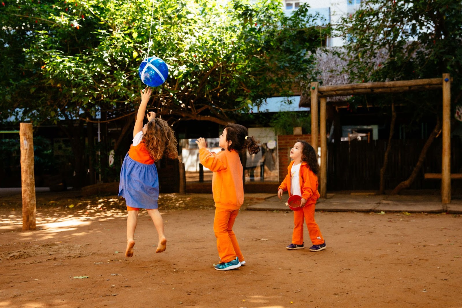 A imagem mostra três crianças jogando bola em um quintal arborizado.
