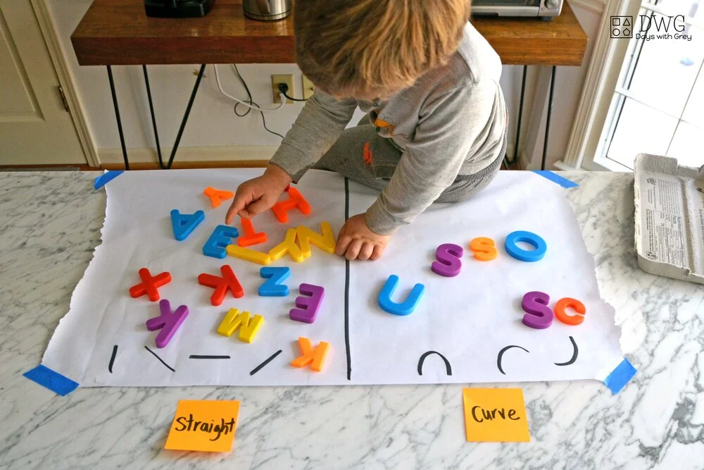 A young boy at a table is sorting colorful magnetic letters into two different piles. One pile is labeled 'straight' and has letters underneath like X, K, M, Z, E, A, Y and the other pile is labeled 'curve' and has letters U, S, O, C.