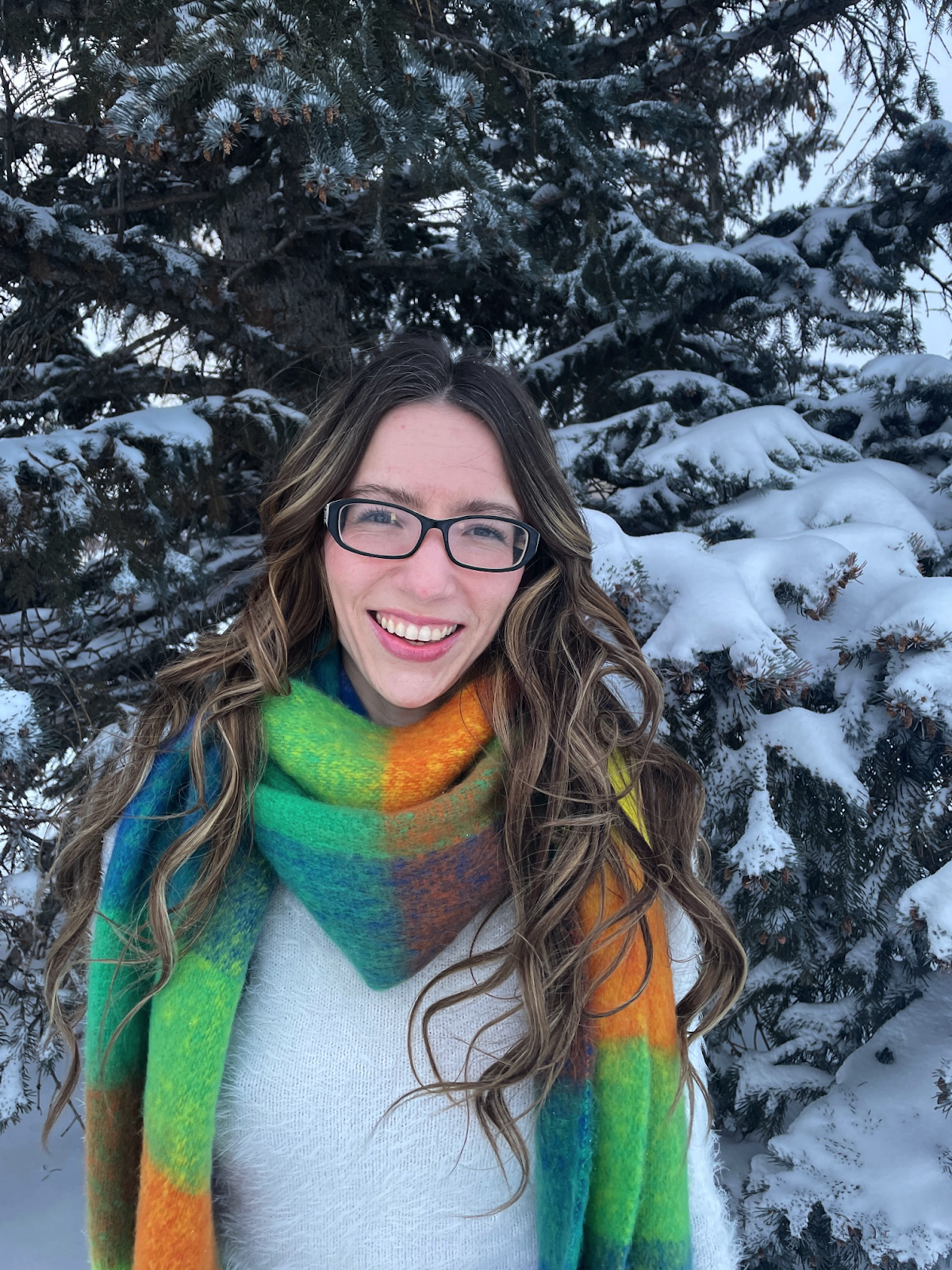 A person wearing glasses and a scarf standing in front of snow covered treesDescription automatically generated with medium confidence